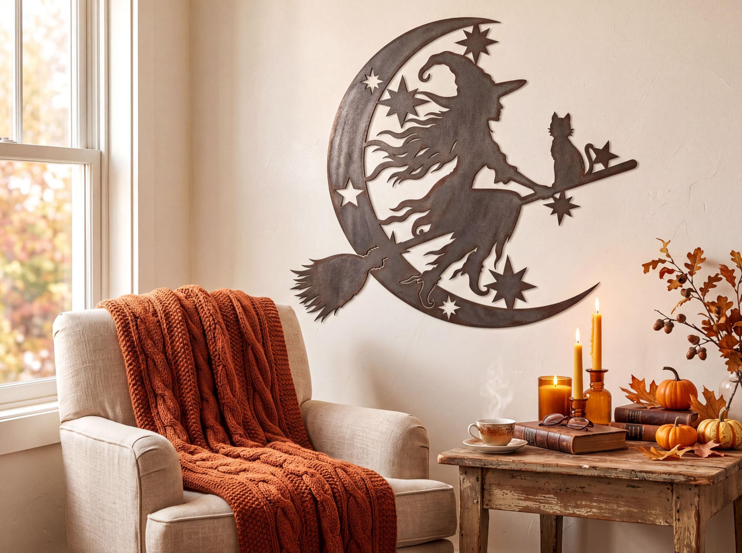 a cozy living room with a large metal crescent moon and stars wall art depicting a witch on a broomstick. The room is decorated with autumn-themed elements, including a pumpkin centerpiece, a candlelit table, and a stack of books. A beige armchair is positioned in front of the wall art, and a small side table is nearby.