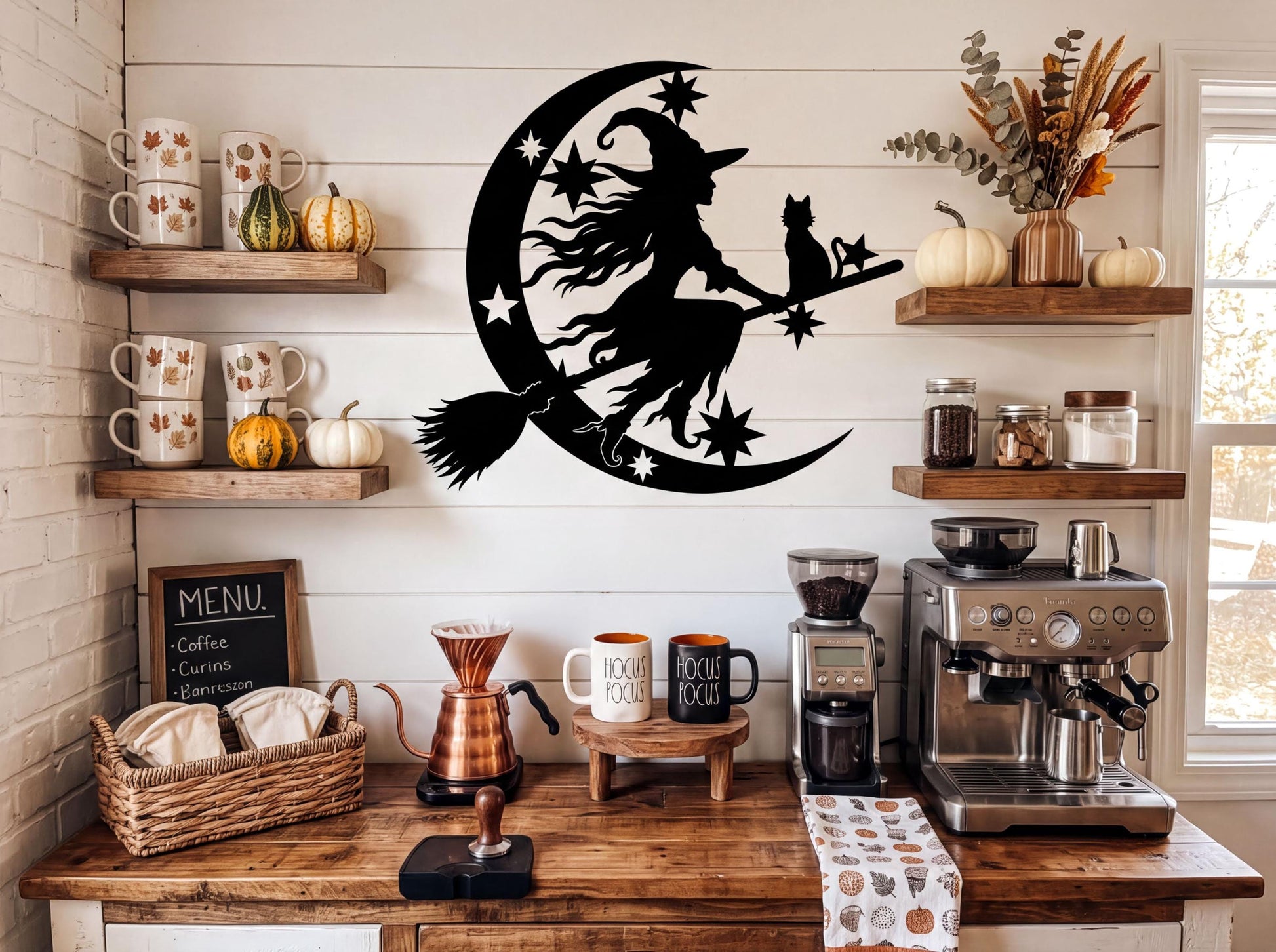 a kitchen counter with various coffee-making equipment, including a coffee maker, a French press, and a percolator. Above the counter, there is a wall decoration depicting a witch riding a crescent moon and holding a cat. The counter also has several coffee mugs, a basket, and some decorative items such as pumpkins and a vase.
