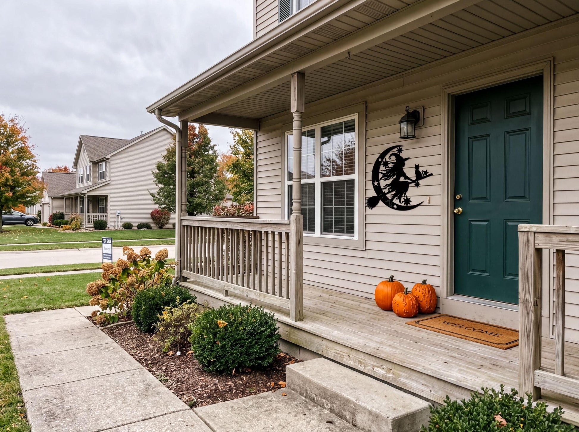 a two-story house with a porch, decorated for Halloween with orange pumpkins and a witch decoration on the front door. The house has a green door and a porch with a railing. The porch is set against a backdrop of trees and a cloudy sky.