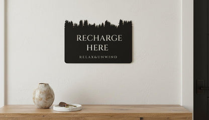 a black sign with white text that reads "RECHARGE HERE" and "RELAX&UNWIND" hanging on a white wall above a wooden table.