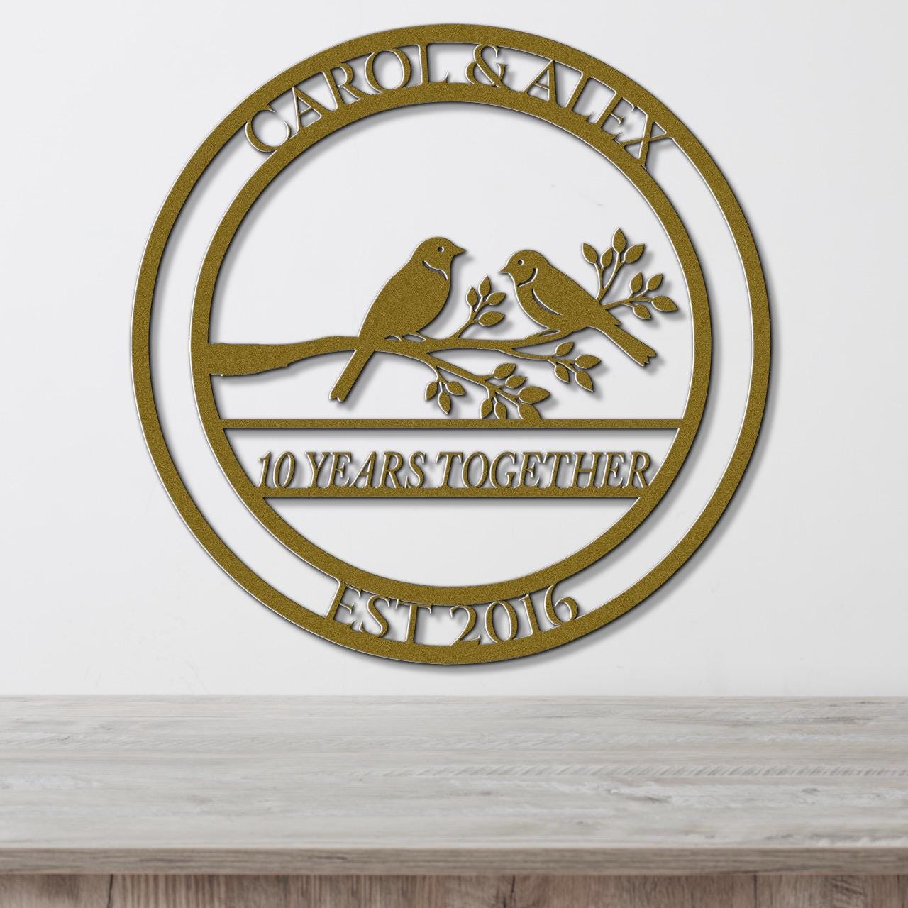 A gold-colored metal sign with the text "CAROL & ALEX 10 YEARS TOGETHER EST 2016" and a graphic of two birds perched on a branch.