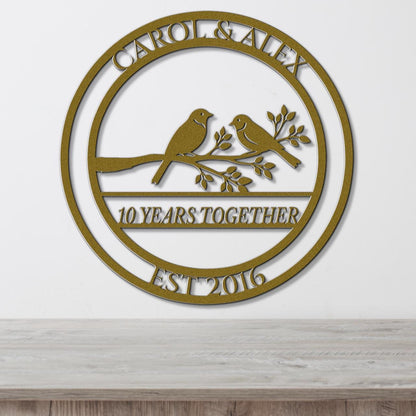 A gold-colored metal sign with the text "CAROL & ALEX 10 YEARS TOGETHER EST 2016" and a graphic of two birds perched on a branch.
