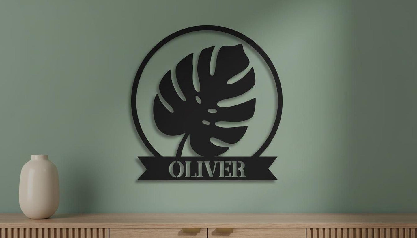 a black metal wall art piece with a large leaf design and the name "OLIVER" below it, mounted on a green wall above a wooden cabinet.