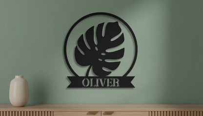 a black metal wall art piece with a large leaf design and the name "OLIVER" below it, mounted on a green wall above a wooden cabinet.