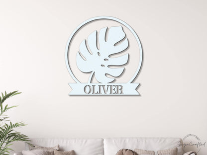a white wall with a circular metal sign that displays a leaf design and the name "OLIVER" below it.