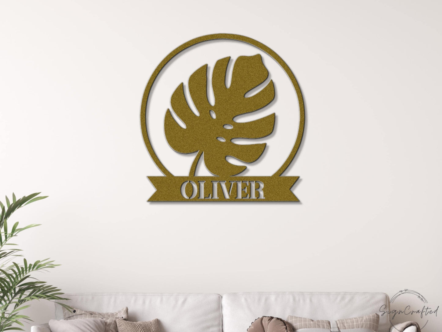 a gold-colored metal wall art piece with a large leaf design and the name "OLIVER" written below it.