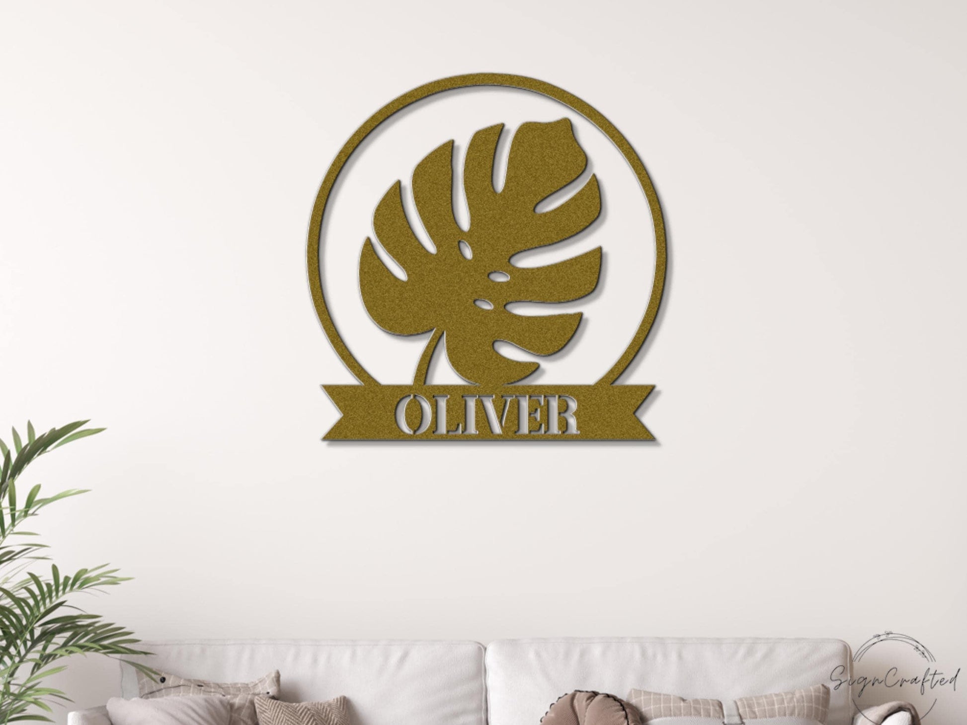 a gold-colored metal wall art piece with a large leaf design and the name "OLIVER" written below it.