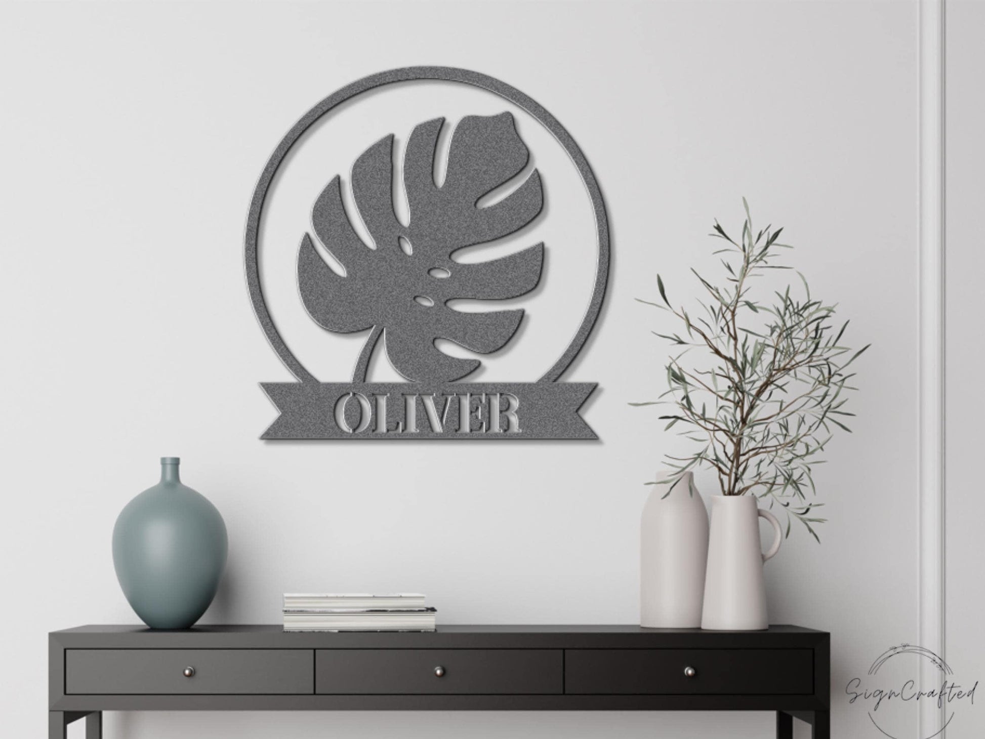 a wall decoration with a large circular metal sign displaying the name "OLIVER" in a bold, capitalized font. Below the sign, there is a black dresser with a vase and a plant on top.