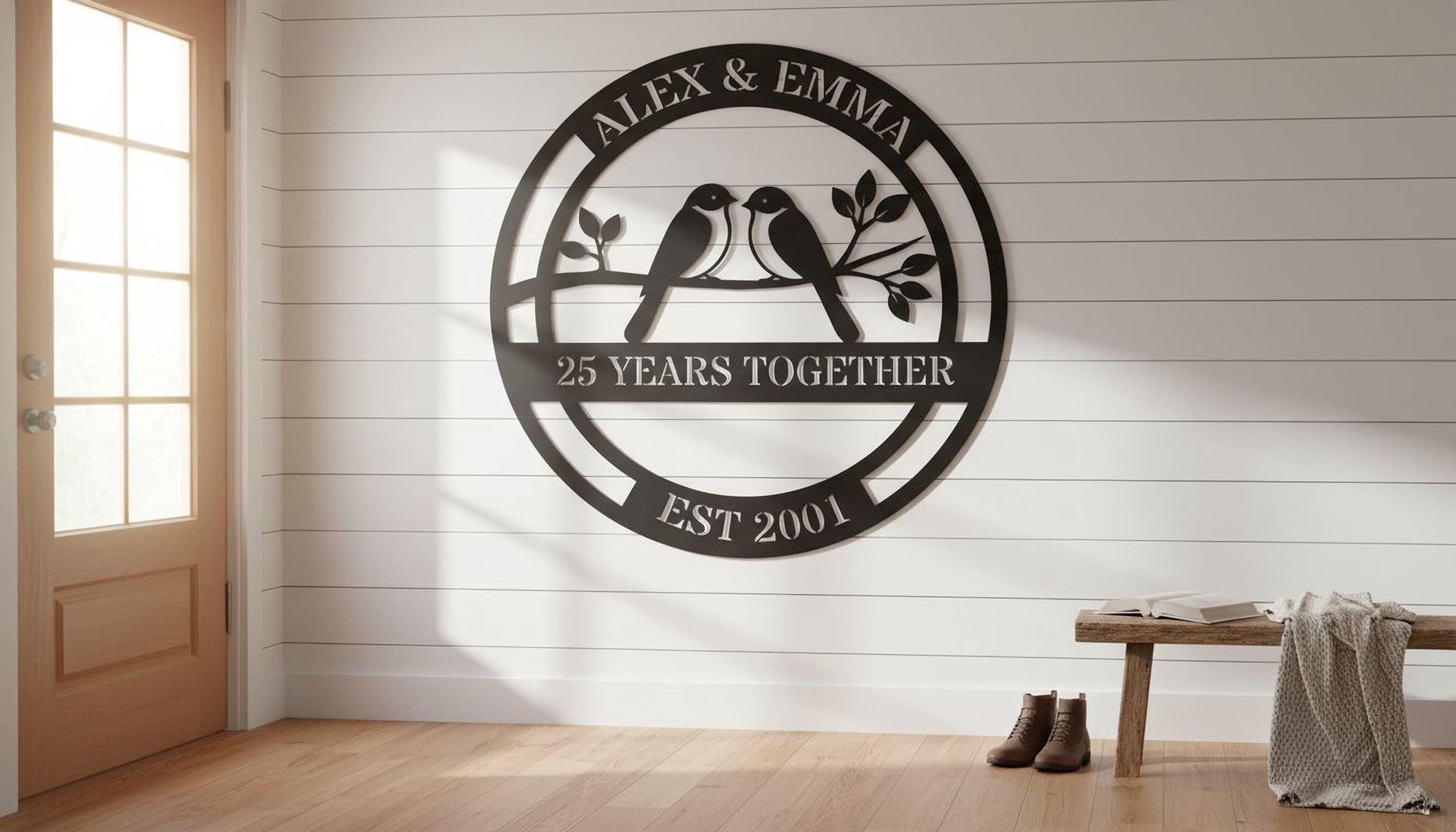 a large black metal sign with the text "Alex & Emma 25 YEARS TOGETHER EST 2001" and a graphic of two birds perched on a branch. The sign is mounted on a white wall, and there is a wooden bench and a pair of boots placed nearby.