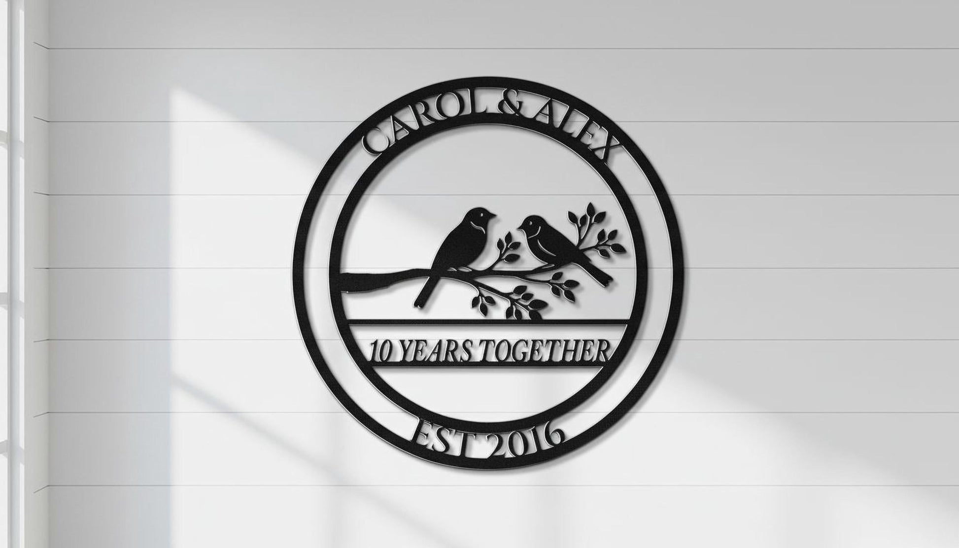 A black and white metal sign with the text "CAROL & ALEX 10 YEARS TOGETHER EST 2016" and a graphic of two birds perched on a branch.