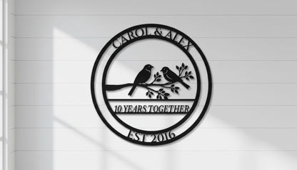 A black and white metal sign with the text "CAROL & ALEX 10 YEARS TOGETHER EST 2016" and a graphic of two birds perched on a branch.