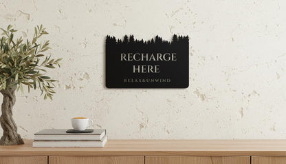 a wooden shelf with a stack of books, a white mug, and a potted plant. Above the shelf, there is a black sign with the text "RECHARGE HERE" and "RELAX & UNWIND" written on it.