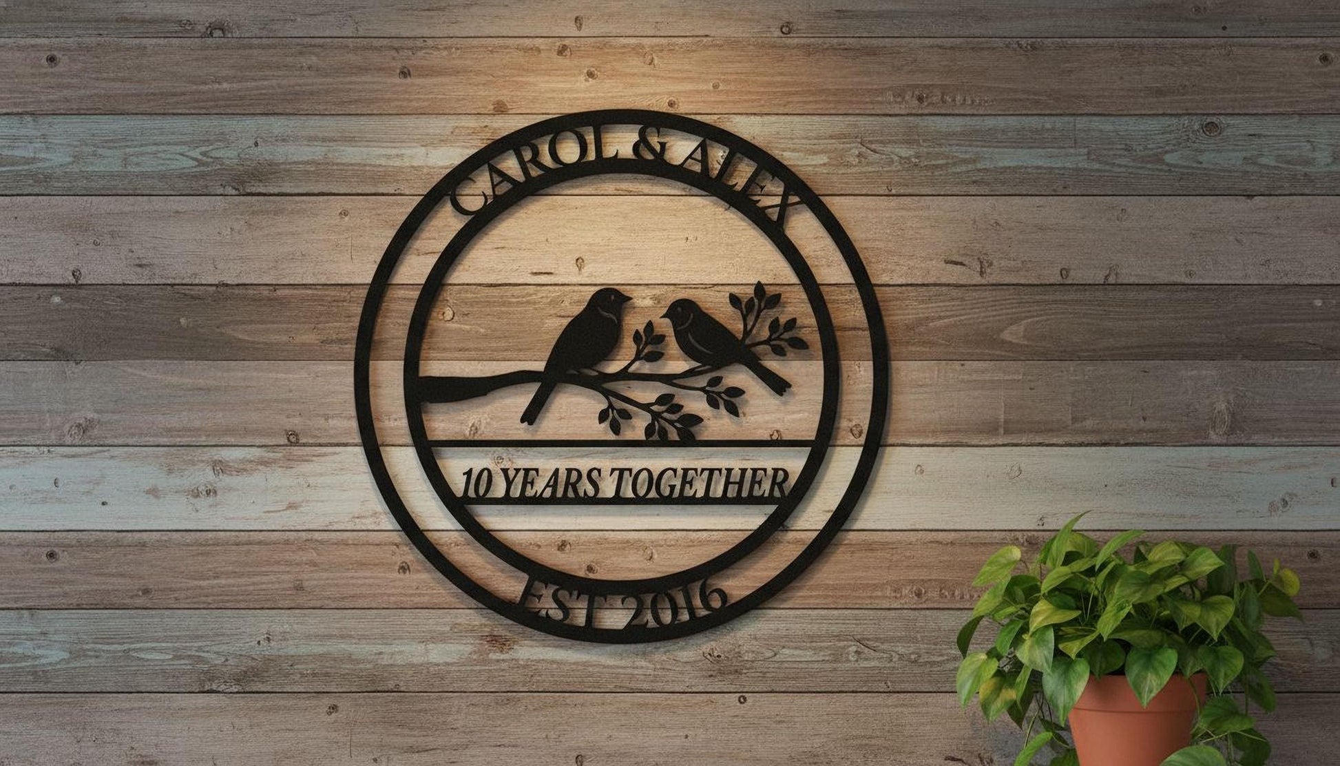 A black metal sign with the text "CAROL & ALEX 10 YEARS TOGETHER EST 2016" hangs on a wooden wall, with a potted plant to the right.