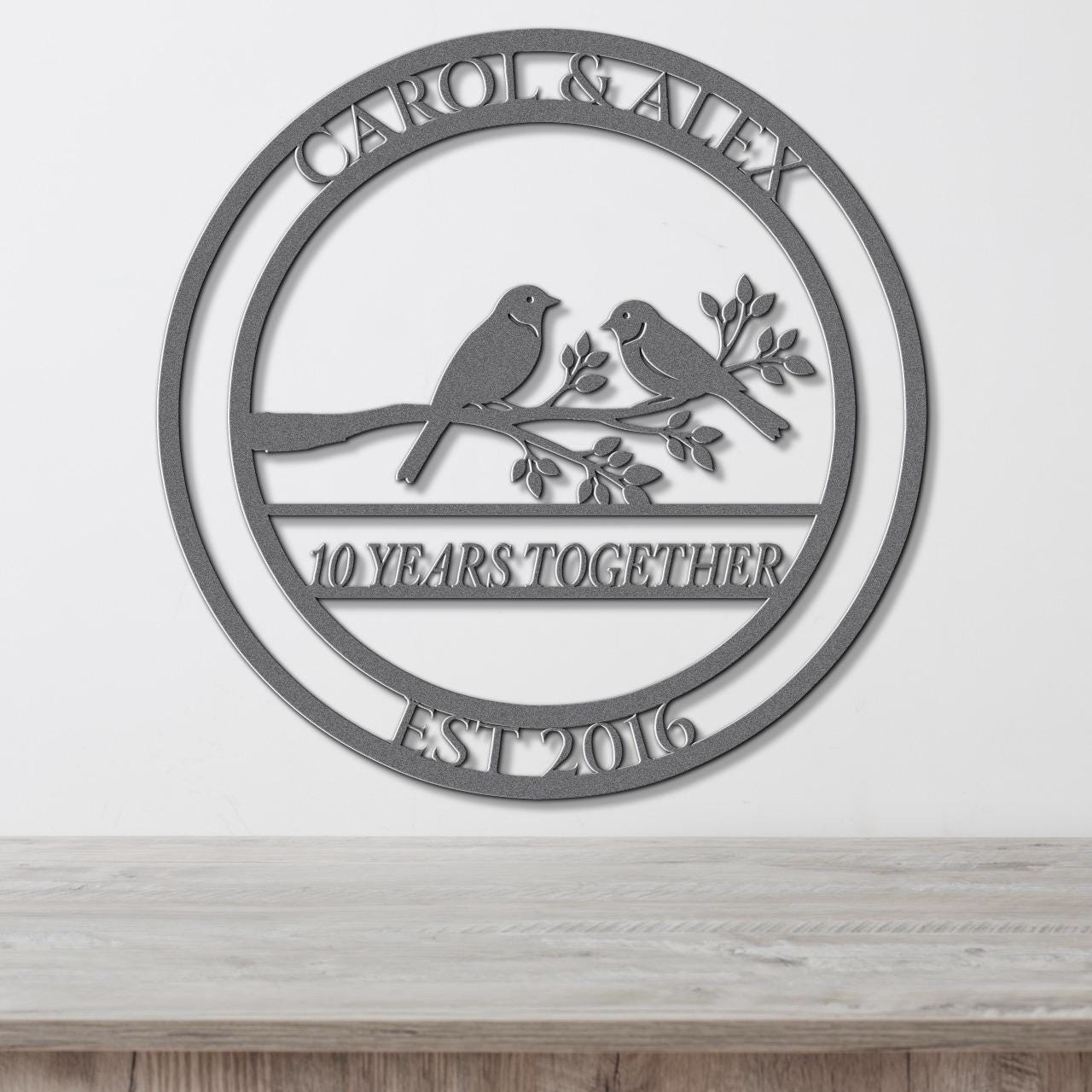 A metal sign with the text "CAROL & ALEX 10 YEARS TOGETHER EST 2016" and a graphic of two birds perched on a branch.