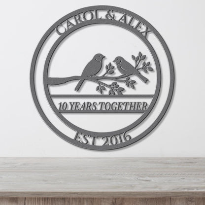 A metal sign with the text "CAROL & ALEX 10 YEARS TOGETHER EST 2016" and a graphic of two birds perched on a branch.