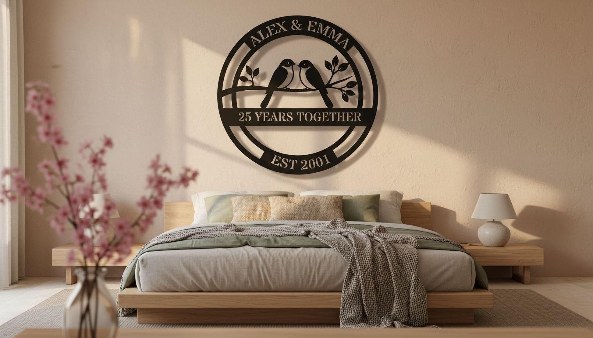 a bedroom with a large metal sign on the wall that reads "ALEX & EMMA 25 YEARS TOGETHER EST 2001". The sign is surrounded by a circular frame with a bird silhouette and the text "25 YEARS TOGETHER". The bedroom has a bed with a gray blanket and a nightstand with a lamp. There is also a vase of pink flowers on the nightstand.
