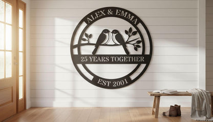 a large black metal sign with the text "ALEX & EMMA 25 YEARS TOGETHER EST 2001" and a graphic of two birds perched on a branch. The sign is mounted on a white wall, and there is a wooden bench and a pair of shoes visible in the foreground.