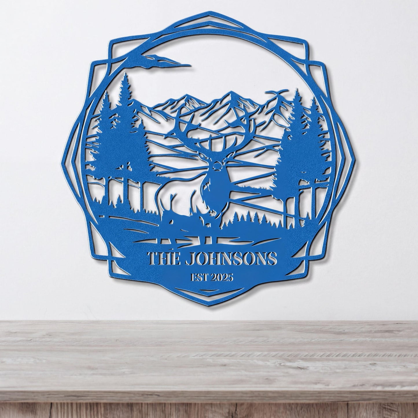 A blue metal wall hanging depicting a deer in a forest landscape with mountains in the background.