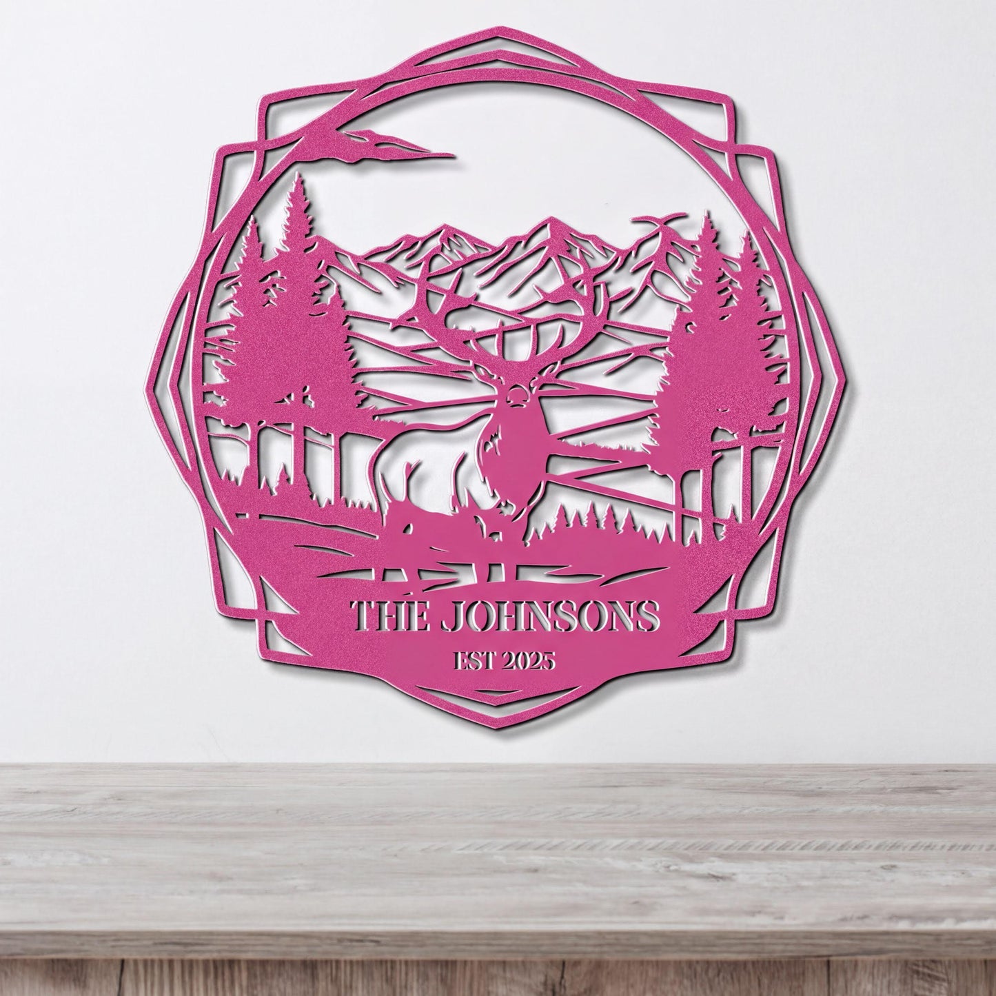A pink metal wall hanging depicting a deer in a forest landscape with mountains in the background.