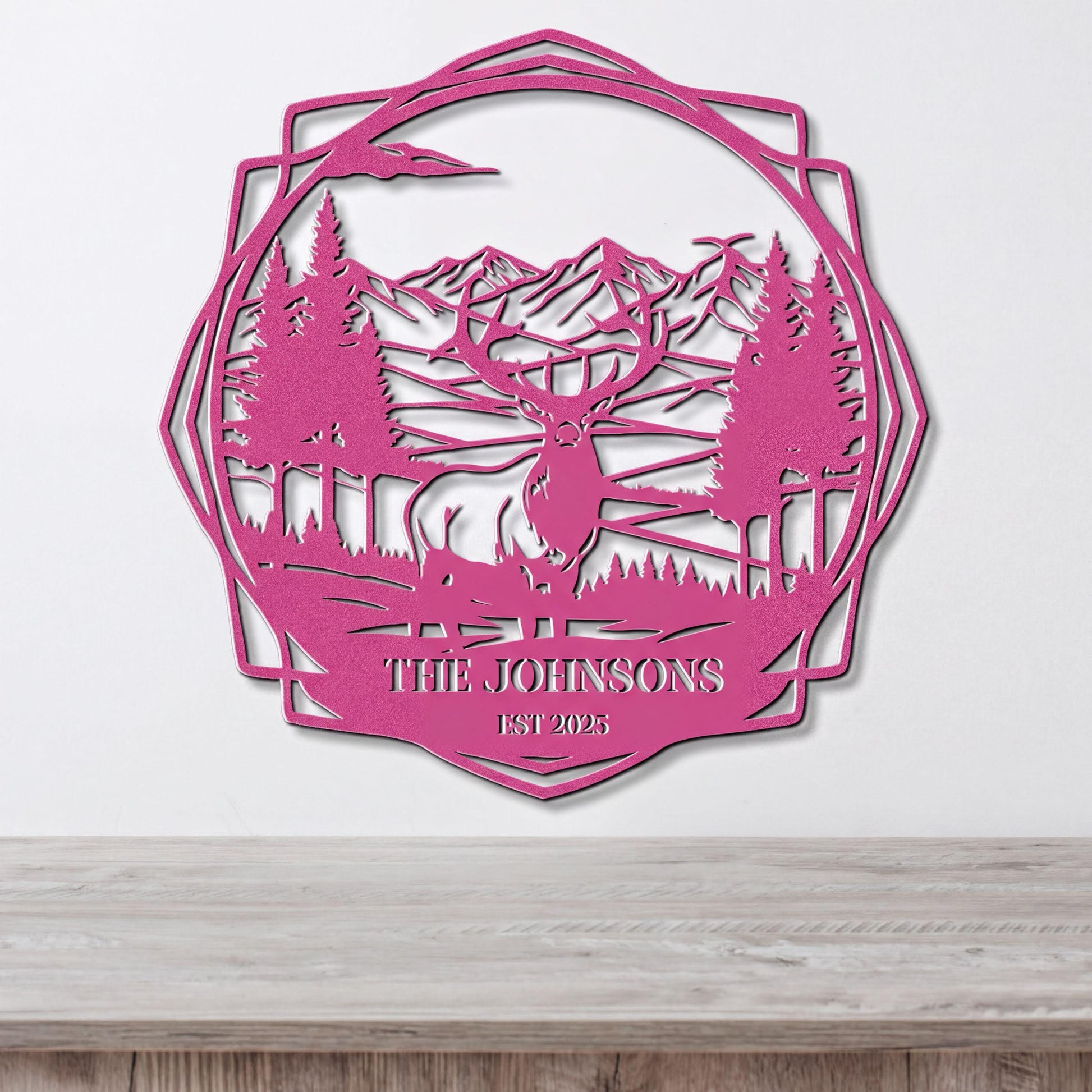 A pink metal wall hanging depicting a deer in a forest landscape with mountains in the background.